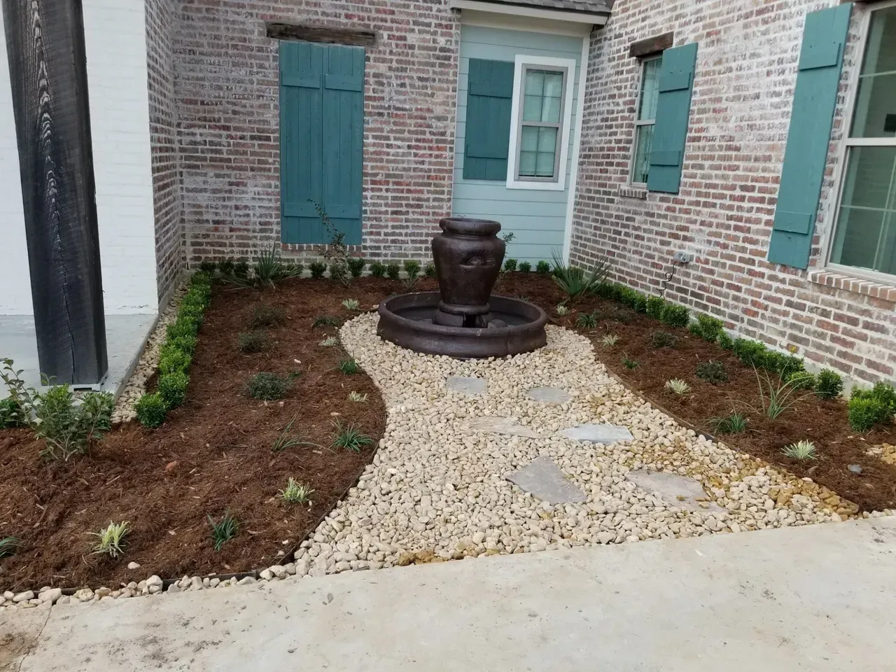 A brick building with a teal-colored fountain and shutters, surrounded by a gravel path and landscaping.