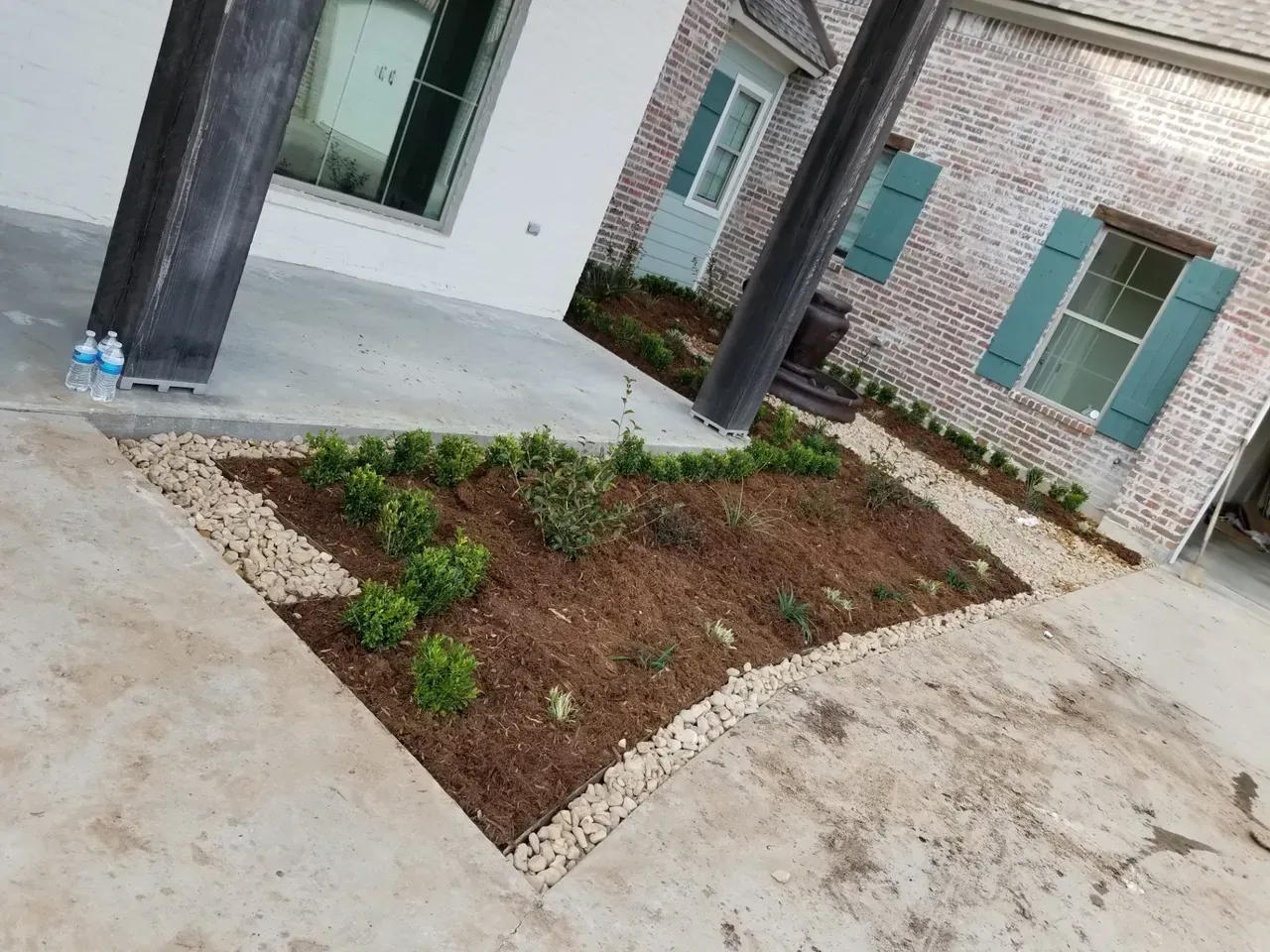 Small landscaped bed with shrubs, edged with rocks, next to a building with brick and turquoise shutters.