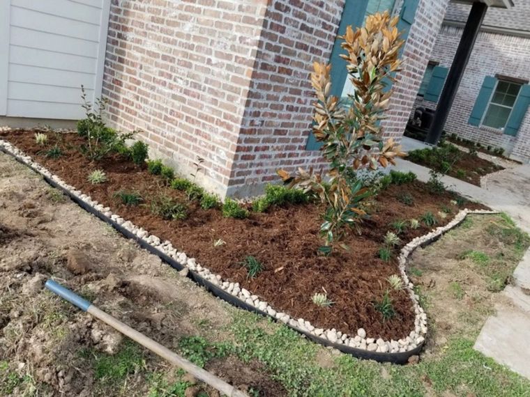 A newly landscaped bed with mulch, rocks, and plants, bordering a brick house. A garden tool rests on the ground.