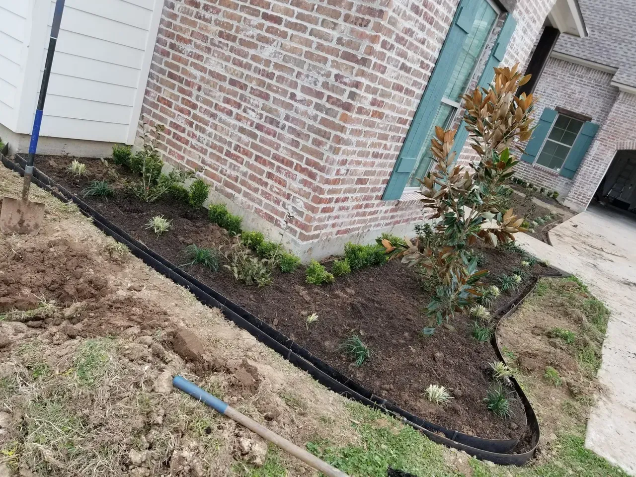 A brick house with a newly planted garden bed edged with black plastic, near a driveway.