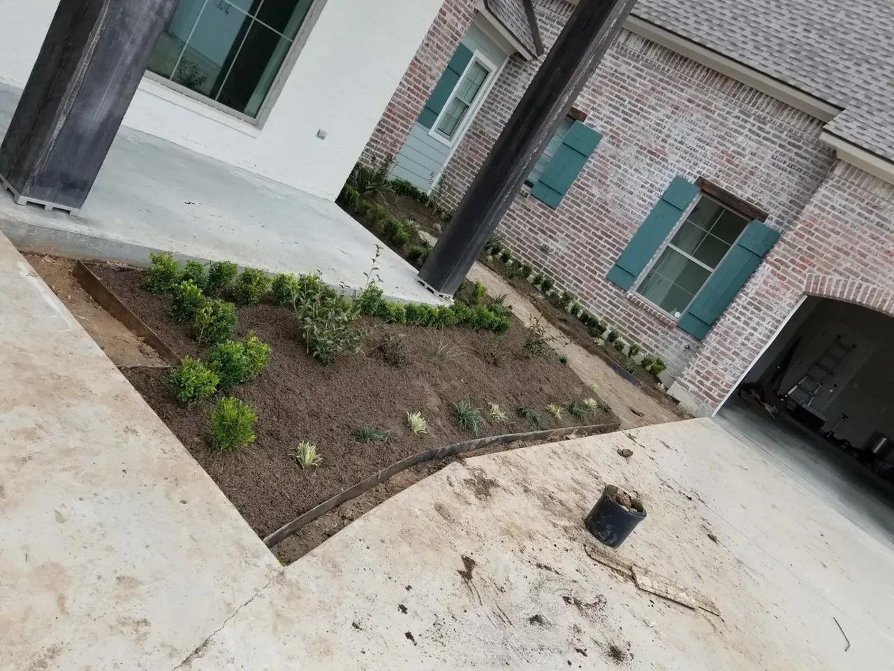 Newly planted garden bed in front of a brick house with green shutters; concrete driveway.