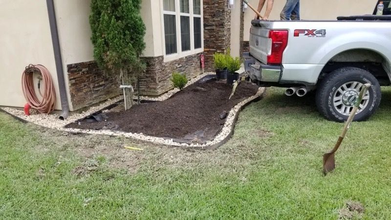 A flower bed with dark mulch and white stone border next to a house and silver truck.