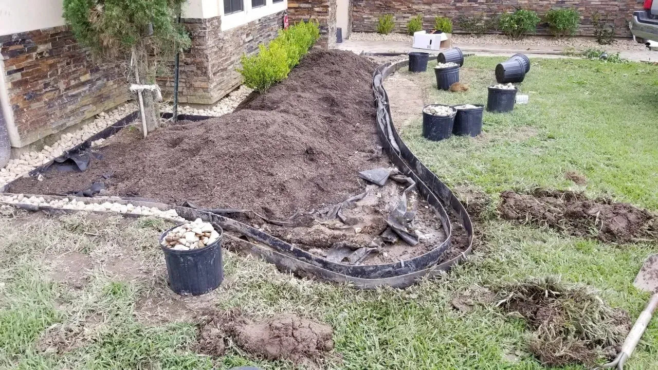A front yard landscape being worked on; dirt, mulch, and pots surround a garden bed edged with black plastic.