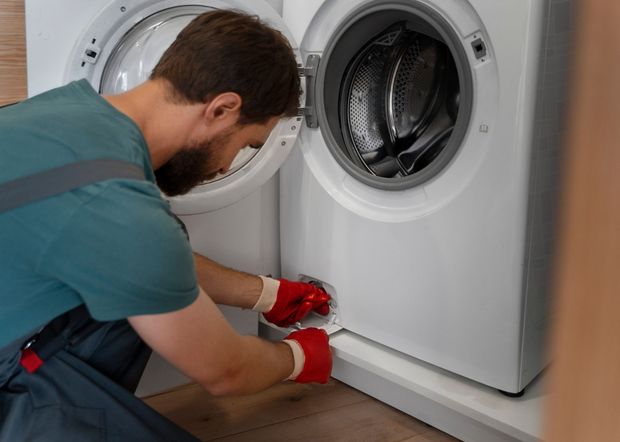 Man in red gloves repairs a white washing machine, door open, kneeling, inside a home.