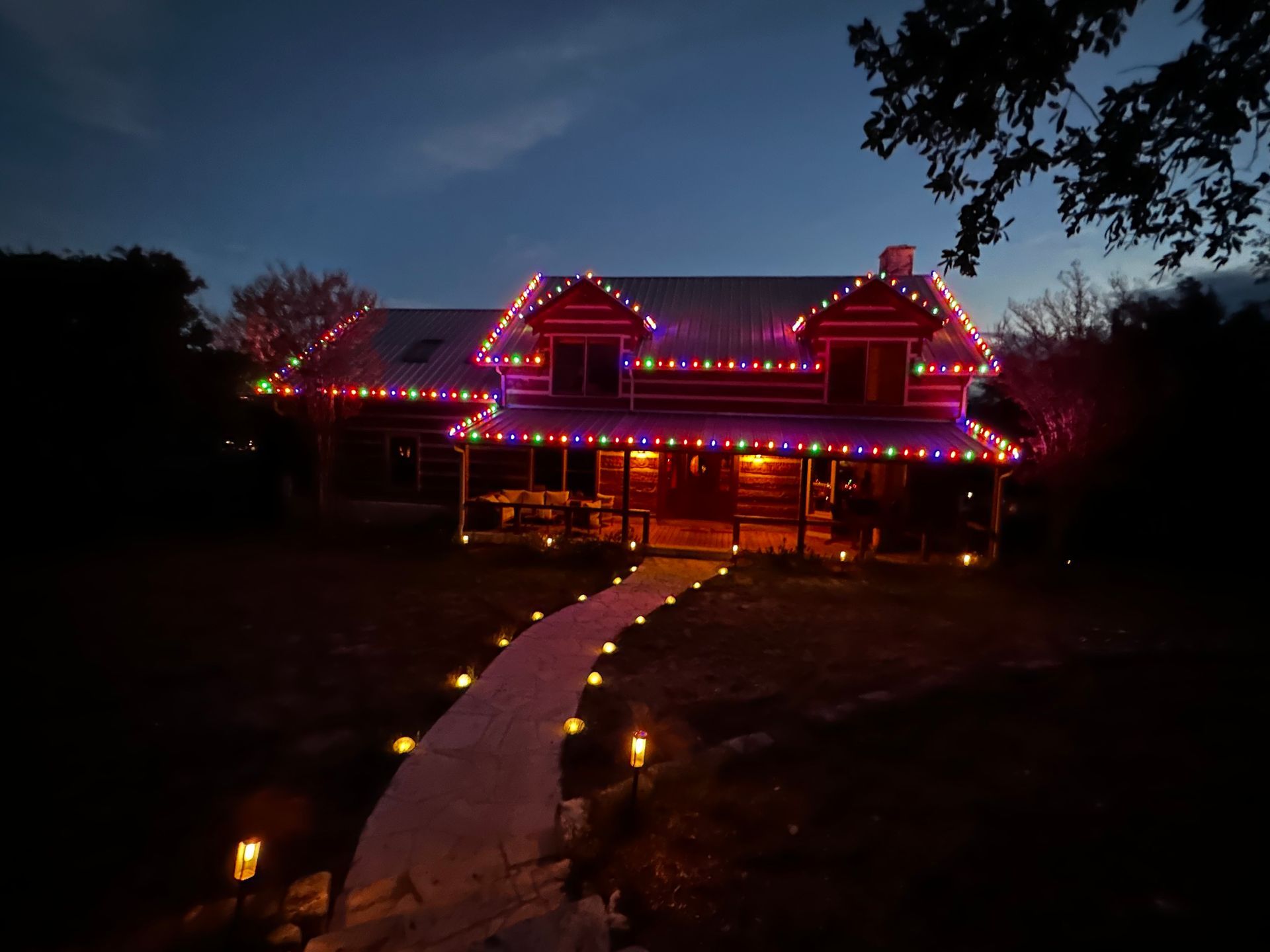 Log cabin at dusk, with colorful Christmas lights outlining the roof. Lit path leads to the porch.