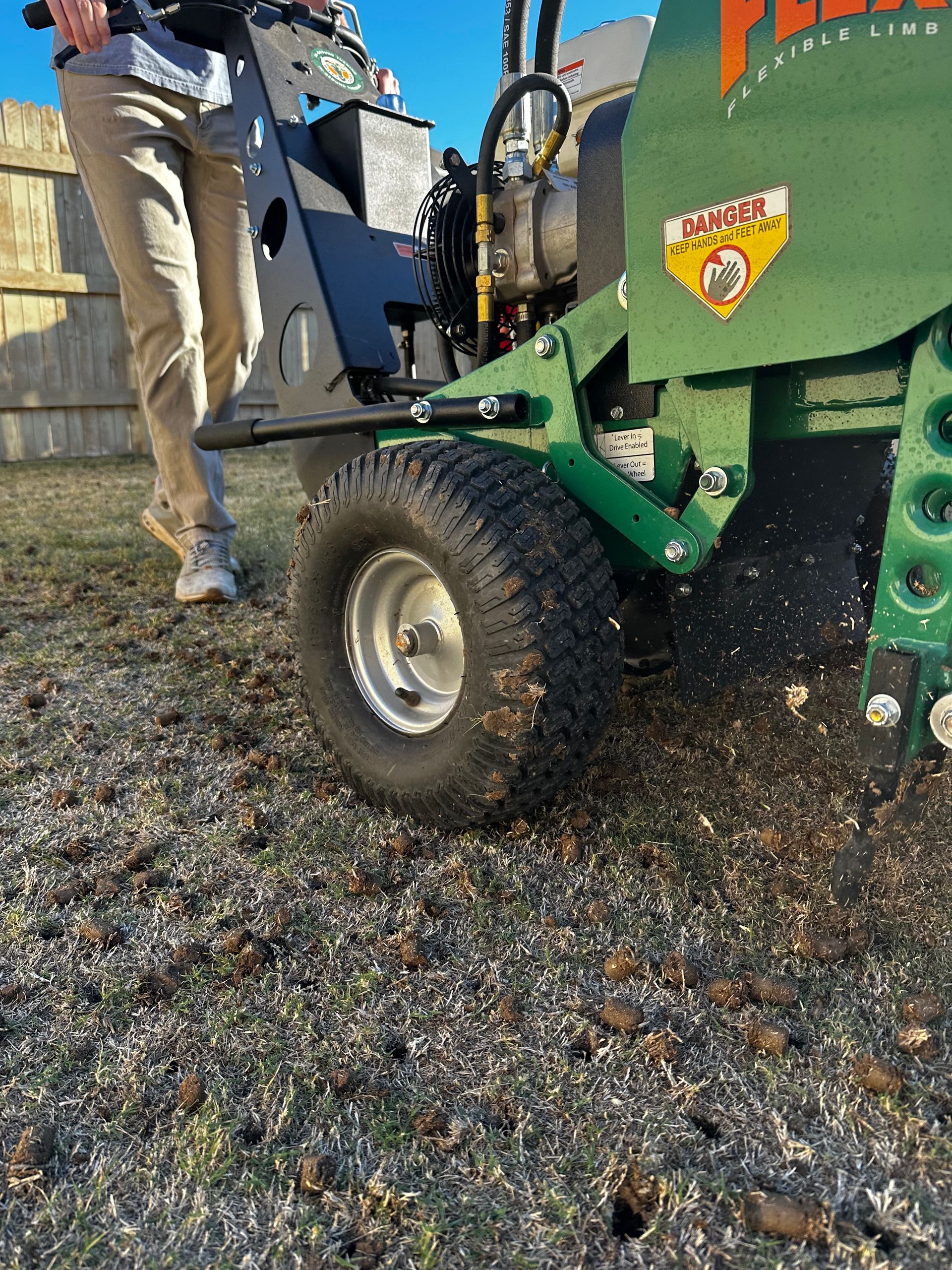 Person beside a green core aerator with a large tire on grass outdoors