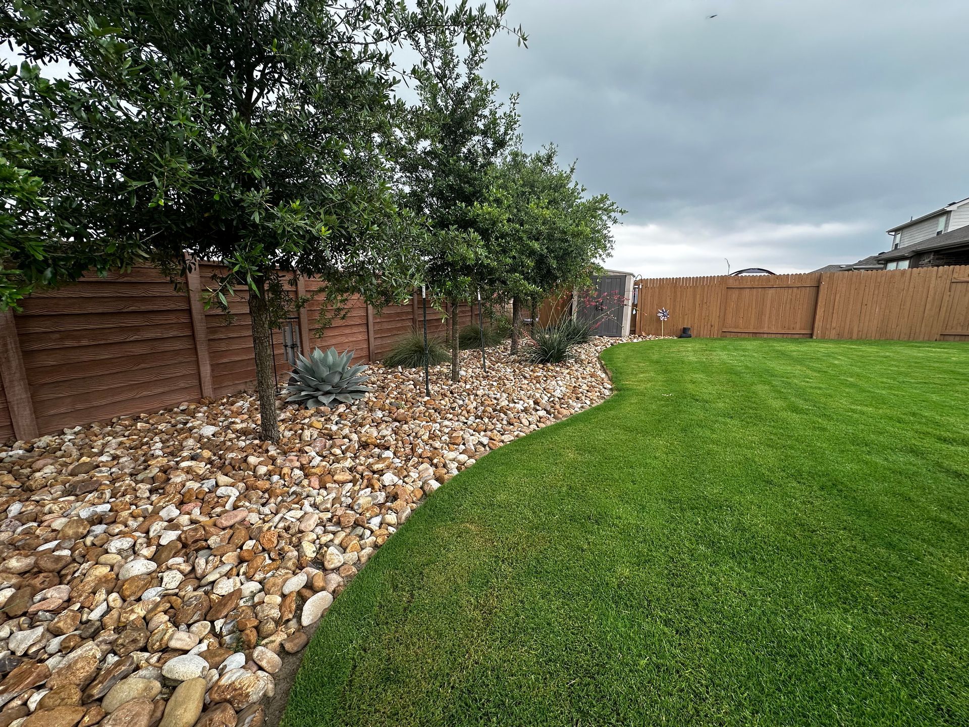 Lush green lawn next to a rock bed with trees, bordered by a wooden fence, under a cloudy sky.