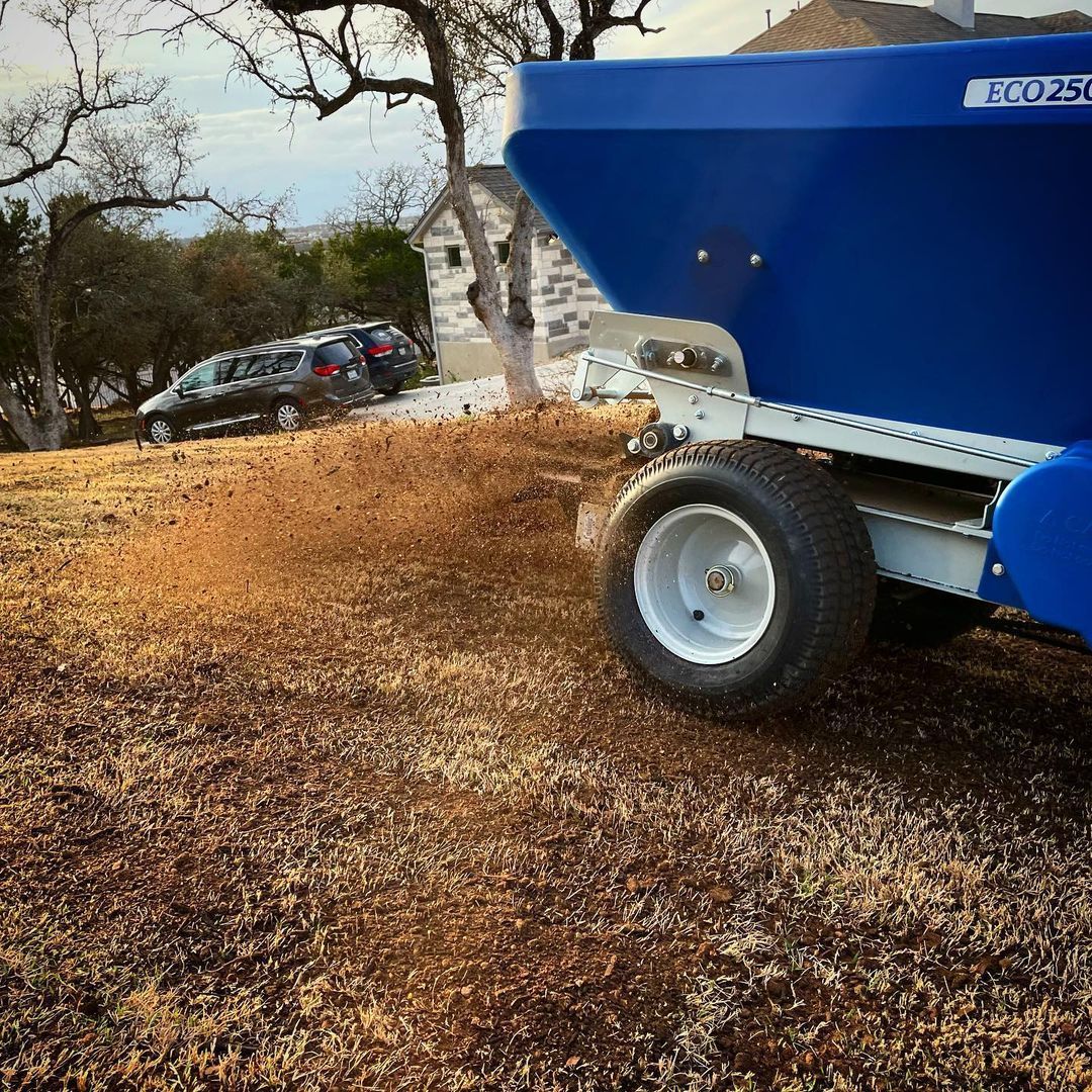 Blue trailer loaded with mulch beside a parked car on a wooded roadside