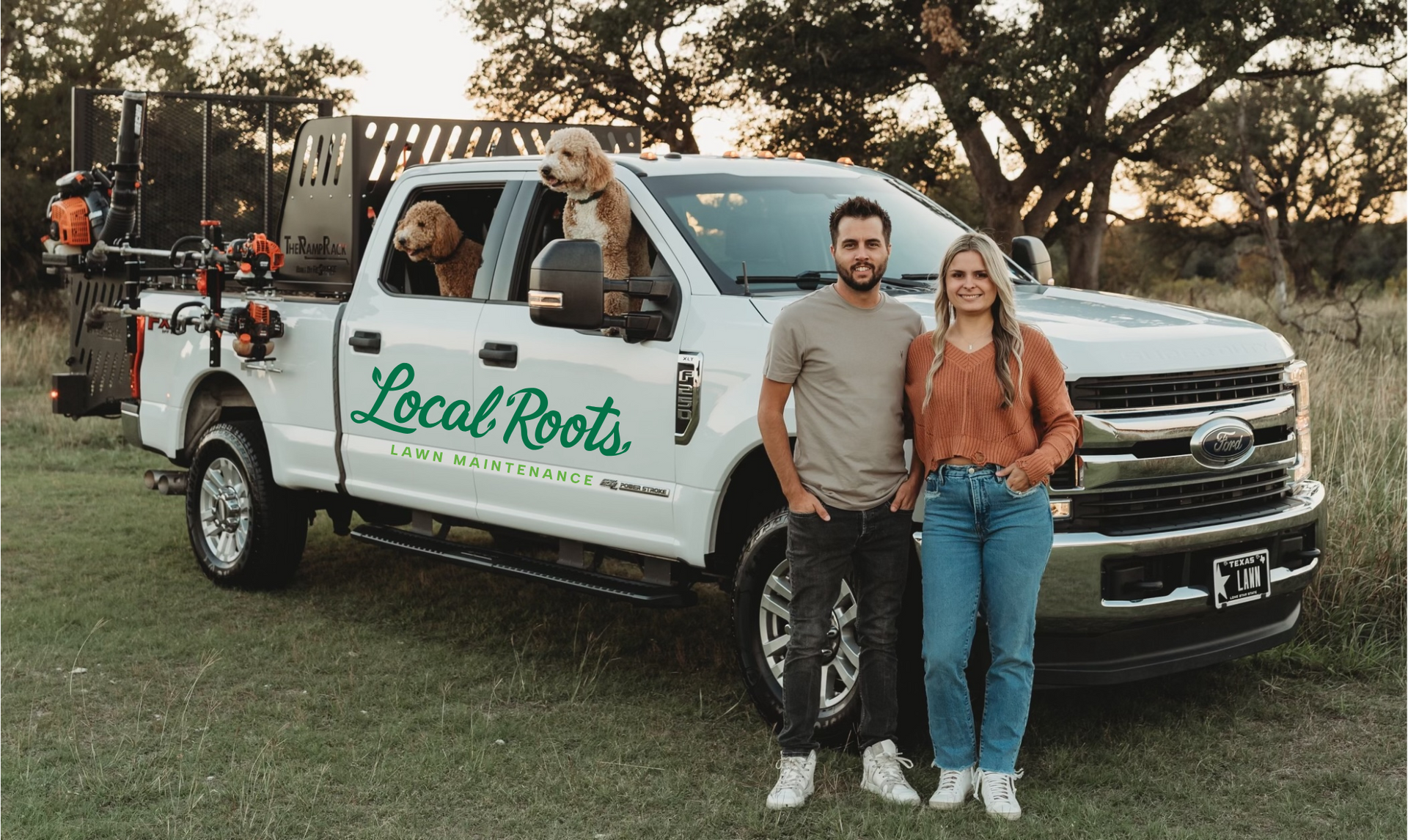 A smiling couple stands next to a white truck of Local Roots Lawn Maintenance with two golden retrievers inside, in a grassy field.