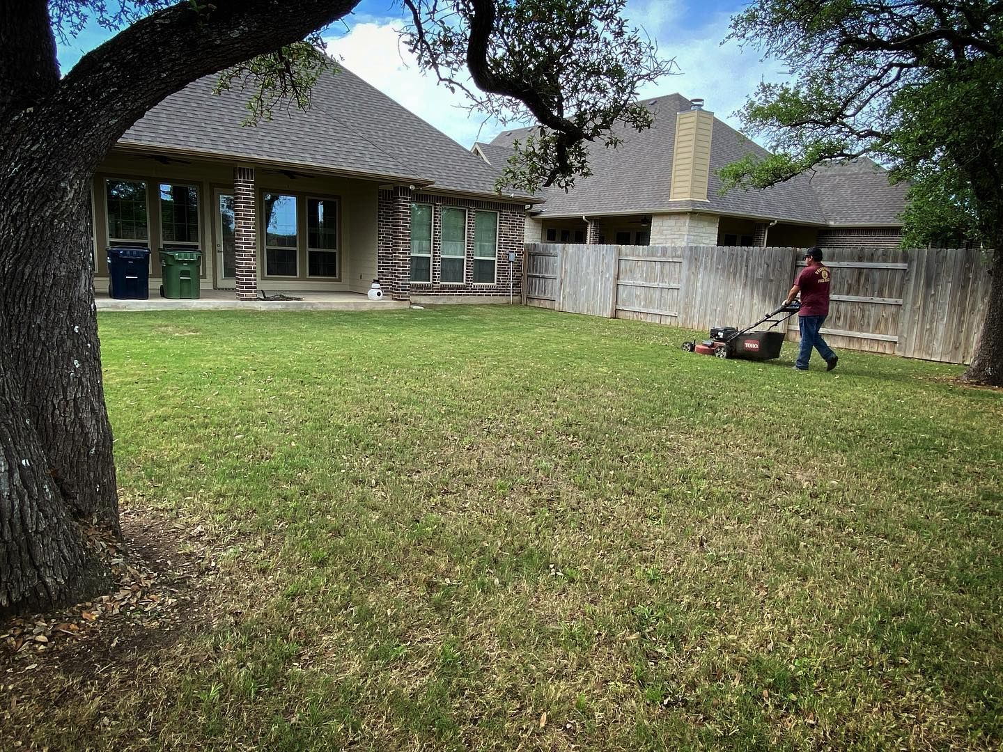 Local roots lawn maintenance worker mowing a green lawn in a backyard, houses and trees visible.