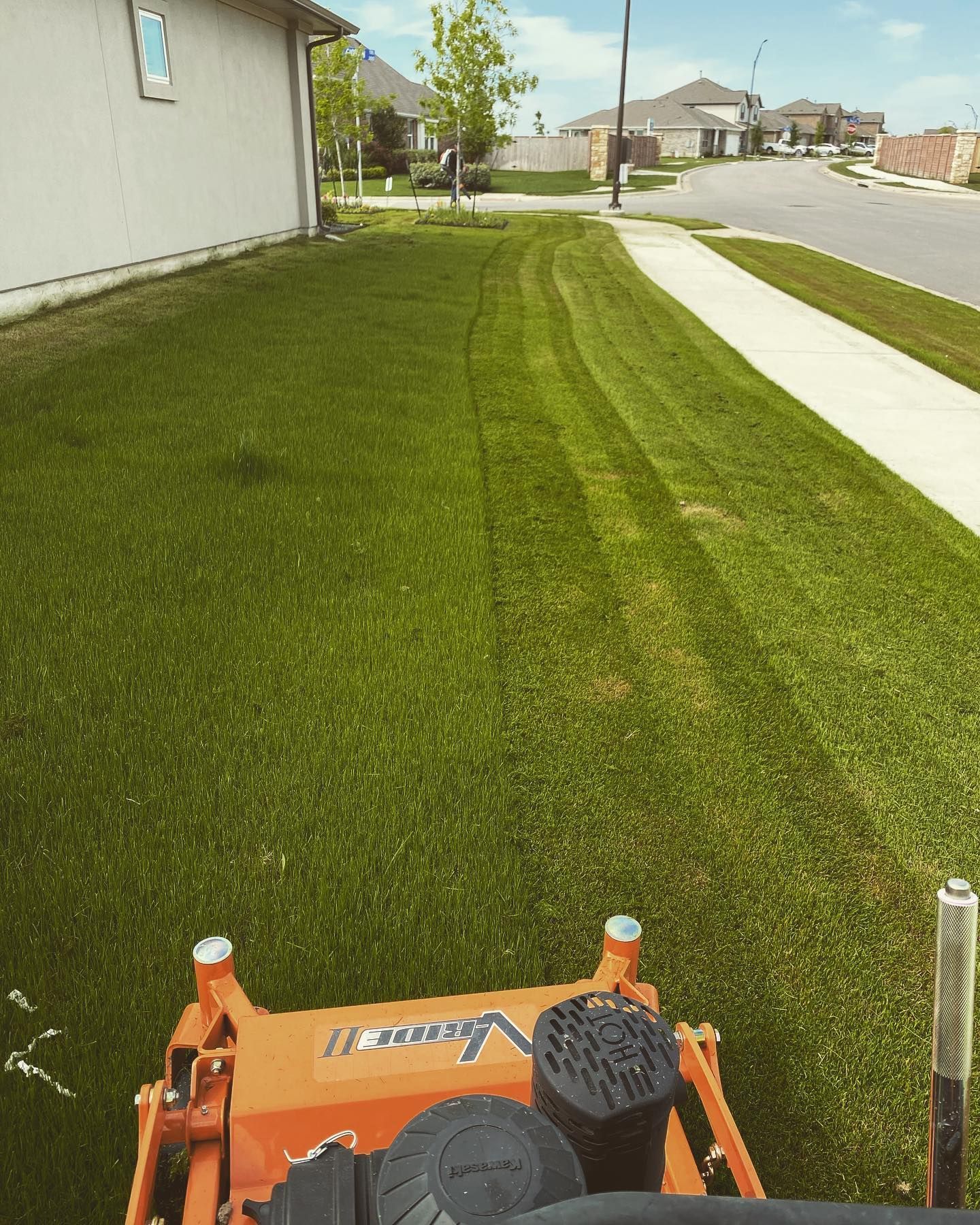 Orange lawn mower cutting grass in a suburban yard. Green grass, blue sky, and white sidewalk.