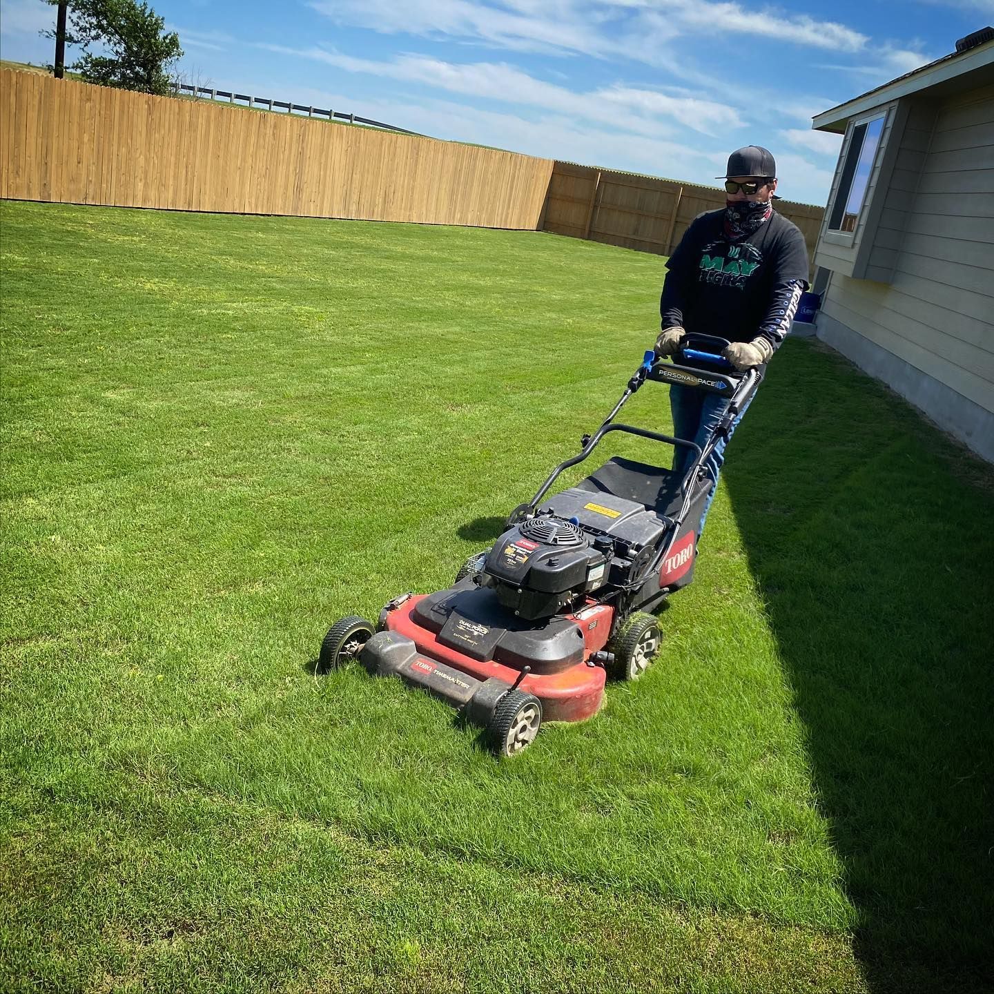 A Local Roots Law Maintenance worker mows a green lawn with a red and black lawnmower next to a house and a wooden fence.
