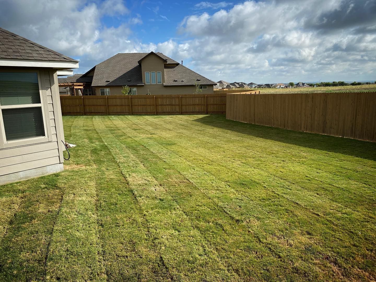 A well-manicured green lawn with a wooden fence, a house and a blue sky with some clouds.