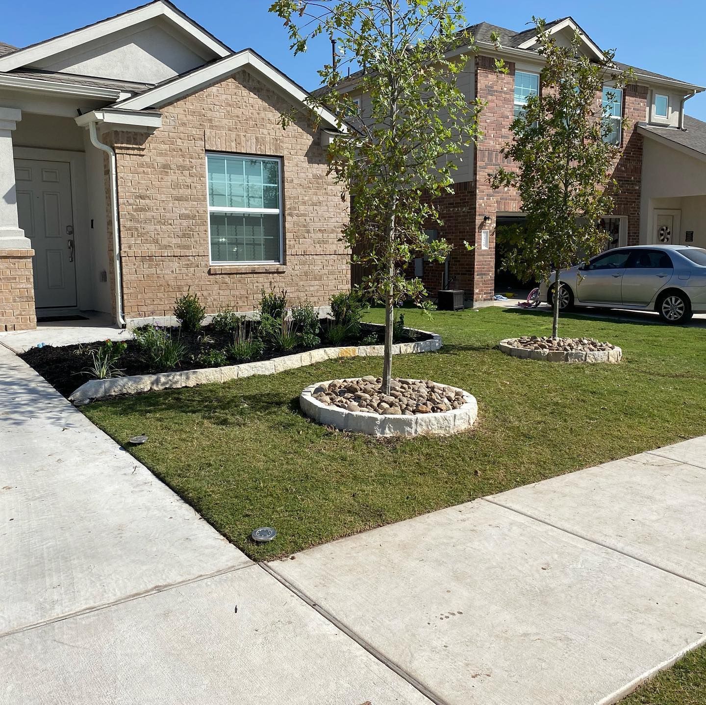 Front yard with brick house, lawn, trees with rock borders, sidewalk, and car.