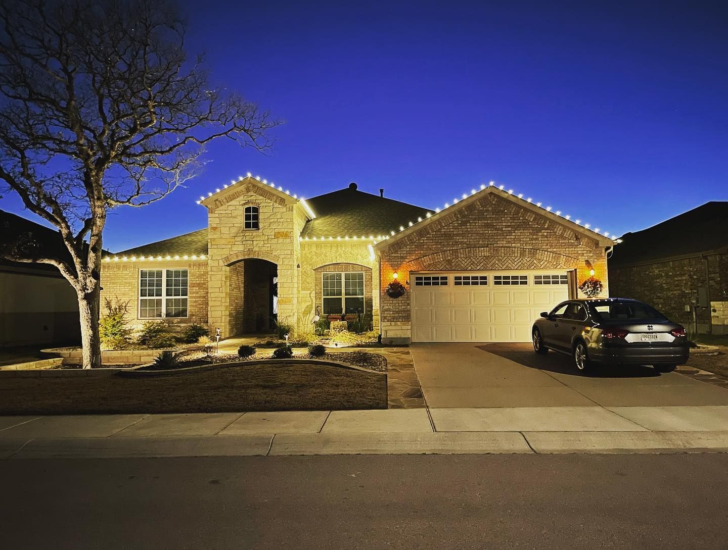 House with Christmas lights, brick facade, driveway with a car, evening sky.