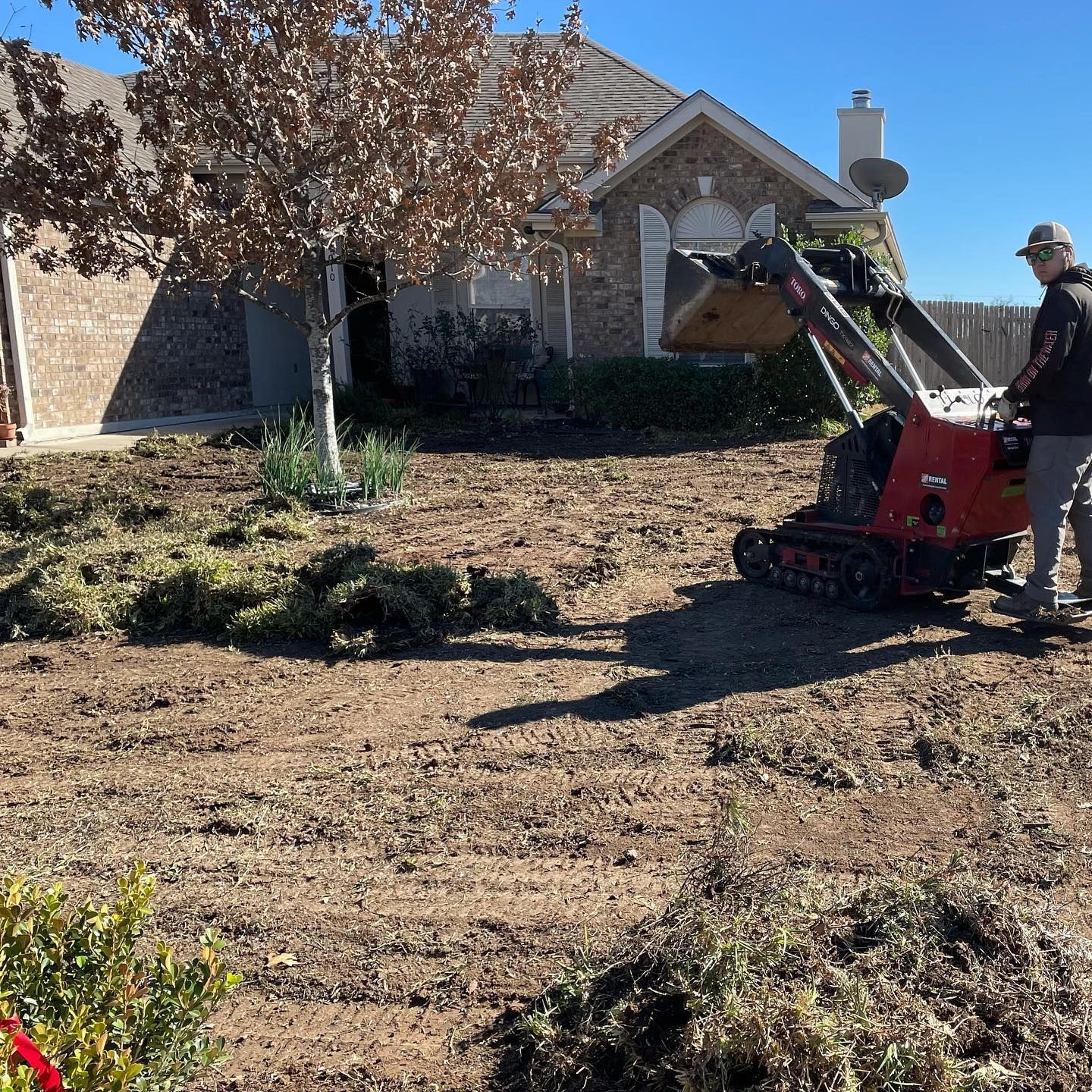 Local roots lawn maintenance worker operating a red mini-excavator in a yard. The ground is dirt and debris near a brick house.
