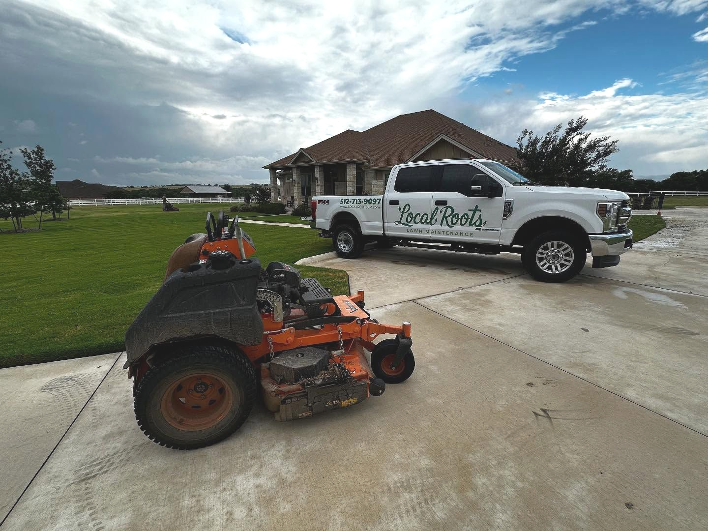 An orange commercial zero-turn lawn mower parked on a driveway next to a white pickup truck labeled