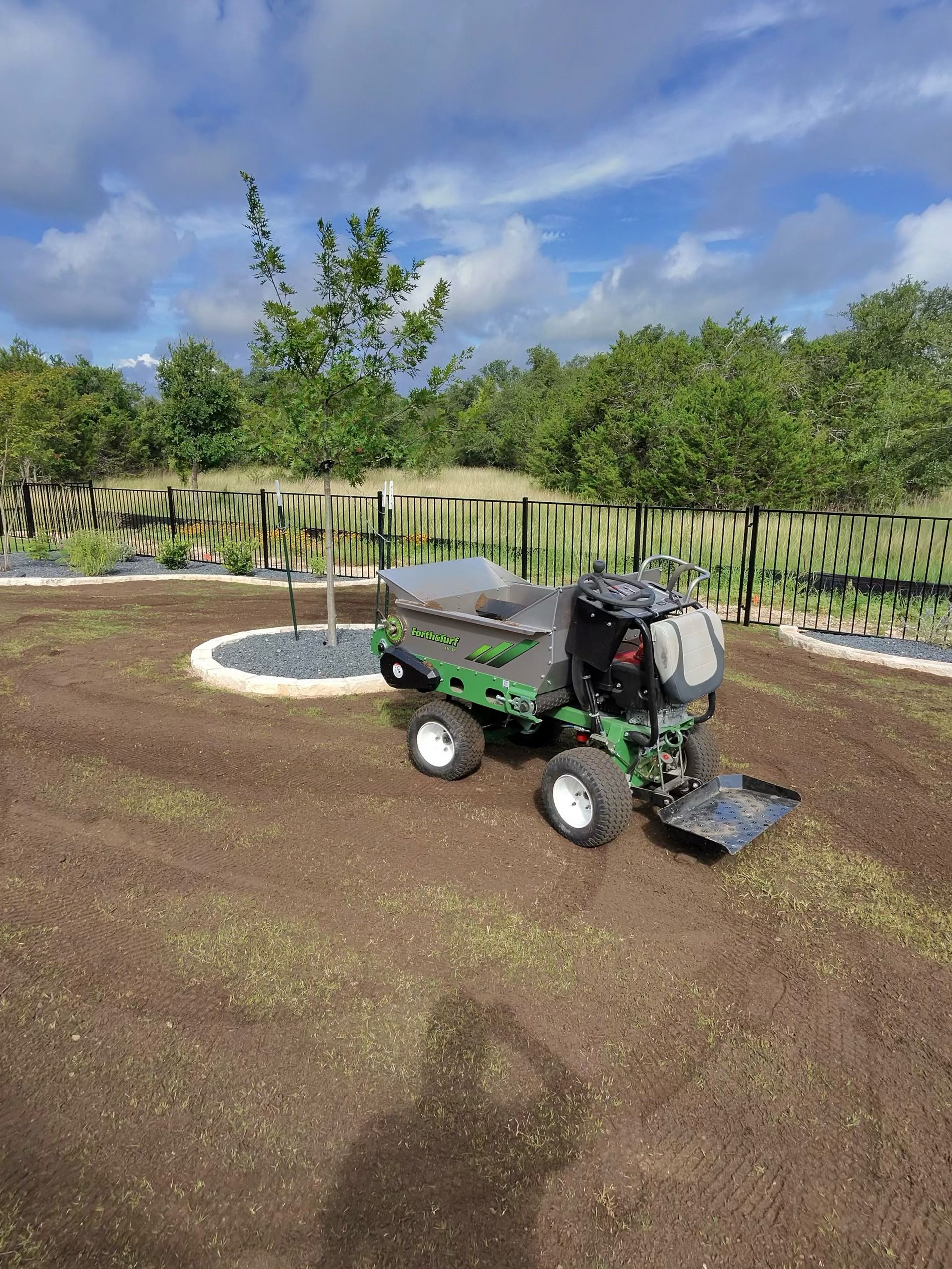 Green motorized dumper on freshly tilled soil in a garden setting. Blue sky, trees in the background.