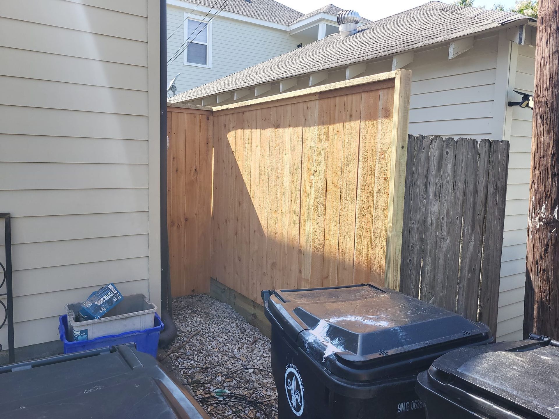 A wooden fence is surrounded by trash cans in a backyard.