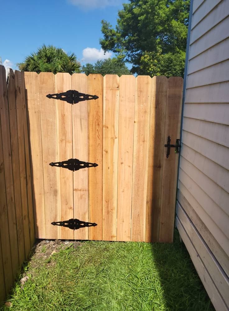 Wooden gate with black hinges and latch in a grassy yard next to a light-colored building.