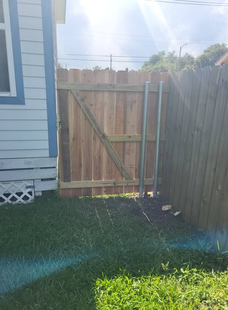 Wooden gate in a wooden fence between a blue house and grassy yard, sunny day.
