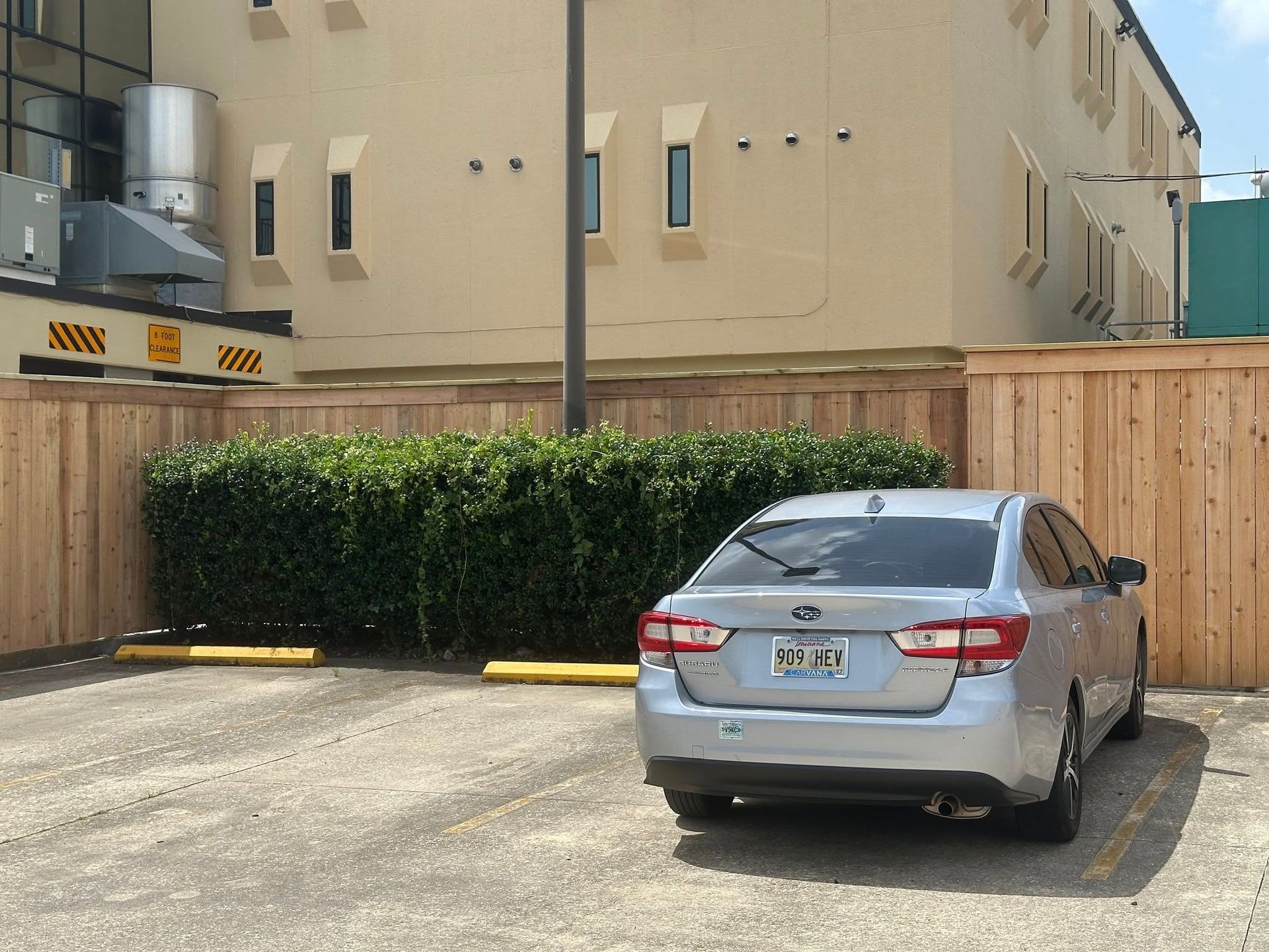 Silver car parked in a lot with wooden fence and bushes in front of a light-colored building.
