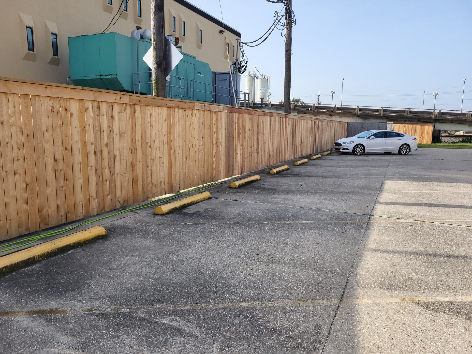 A parking lot with a wooden fence, parking stops, and a white car parked near a building.