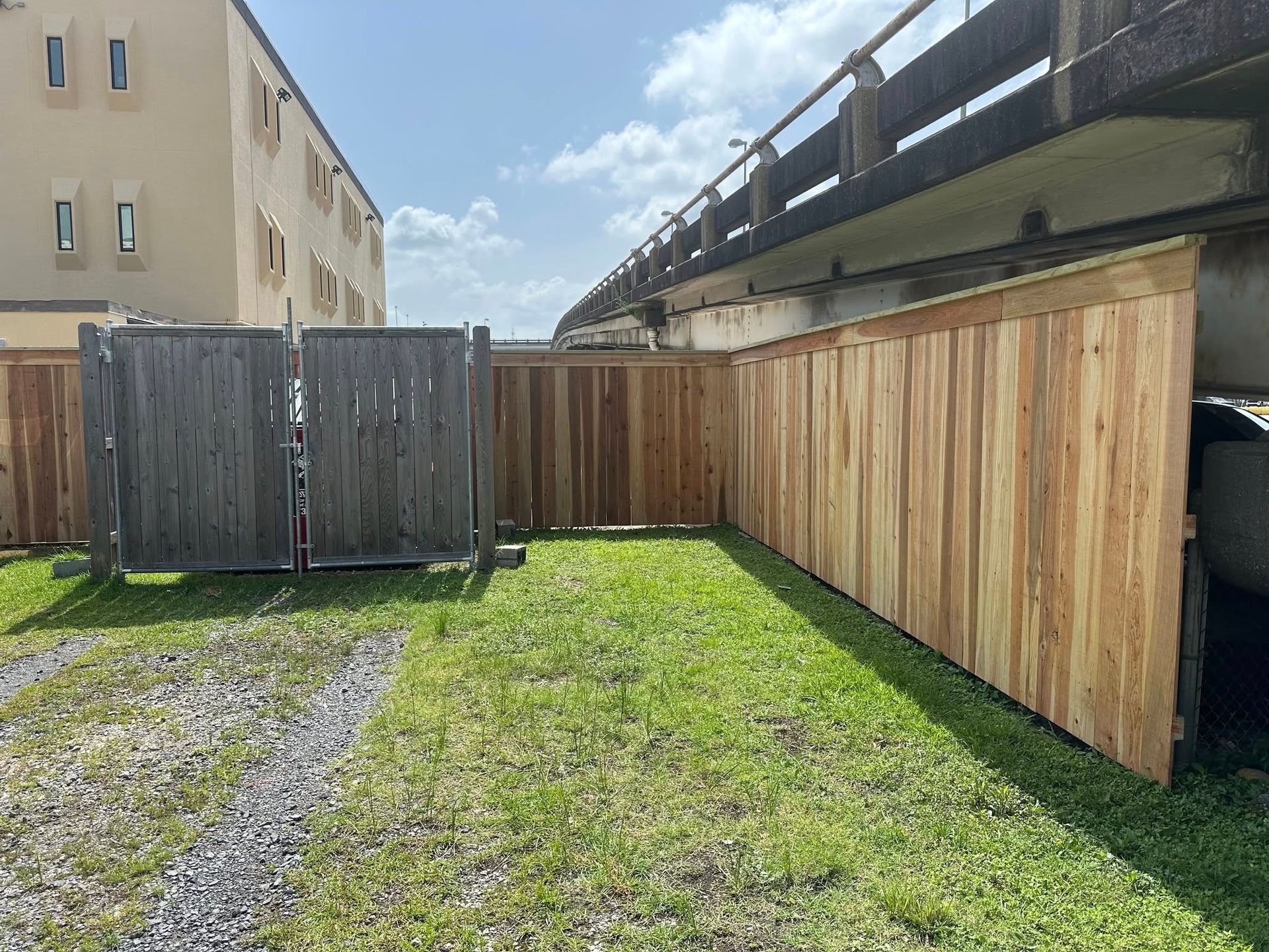 Wooden fences enclose a grassy area under a bridge. Building in the background.
