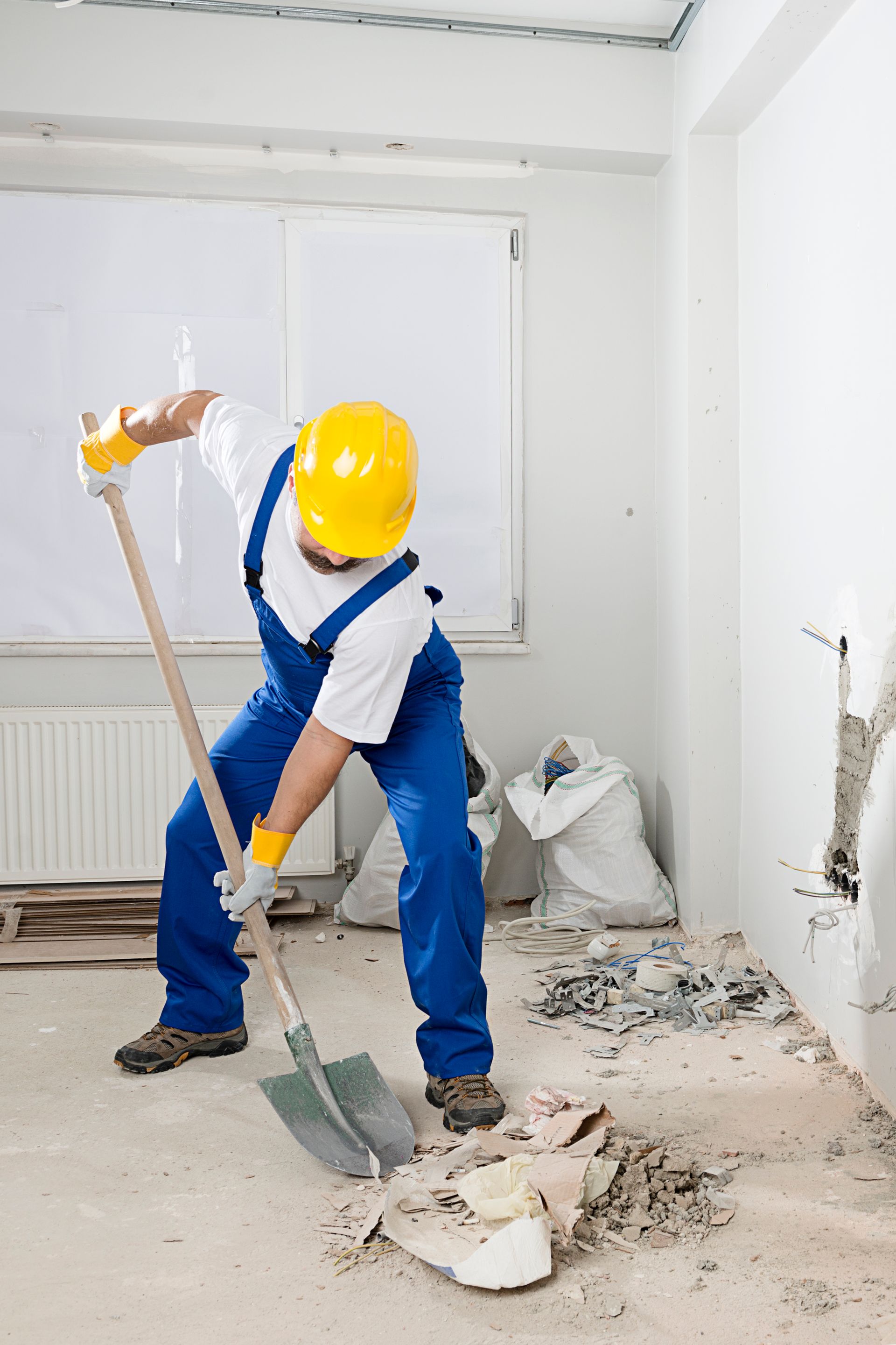 A man wearing a hard hat and overalls is digging with a shovel in a room.