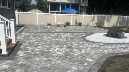 A backyard patio made of grey patterned pavers featuring a white stone-filled fire pit area and a white privacy fence.