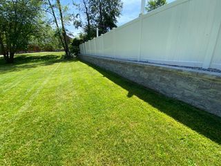 A white vinyl privacy fence sits atop a beige stone retaining wall bordering a vibrant green lawn under a sunny sky.