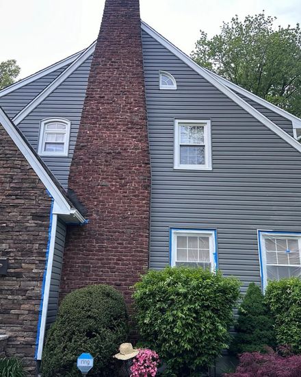 A gray-sided house with a large stone chimney and two stories of windows, with blue painter's tape on the trim.