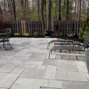 A stone patio with lounge chairs, a table, and a wooden fence, set against a backdrop of trees in an outdoor yard.