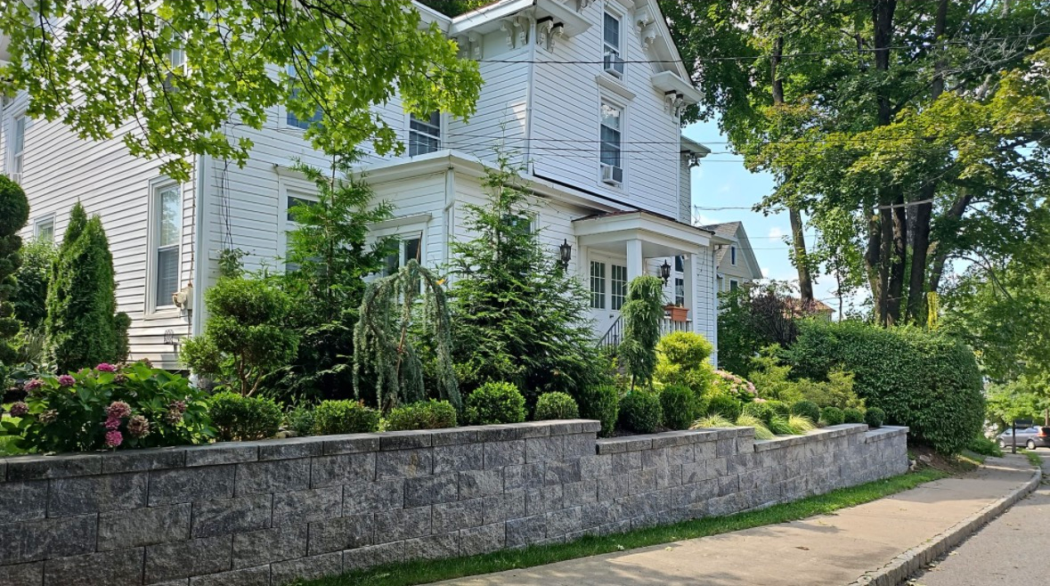 A white, multi-story house with a stone retaining wall and landscaped shrubs in front, situated on a tree-lined street.