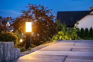 A modern glowing garden bollard light illuminates a stone pathway at dusk, with a dark tree and houses in the background.