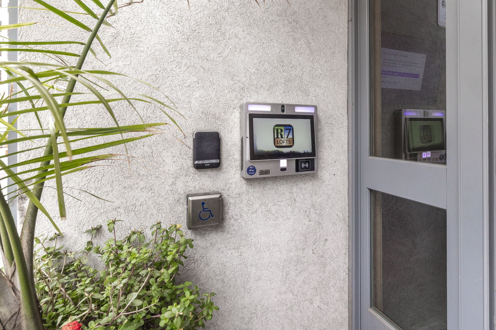 A wall-mounted entry system with screen, card reader, and handicap accessible button, next to a door and plants at R7 Lofts in St. Paul, MN.