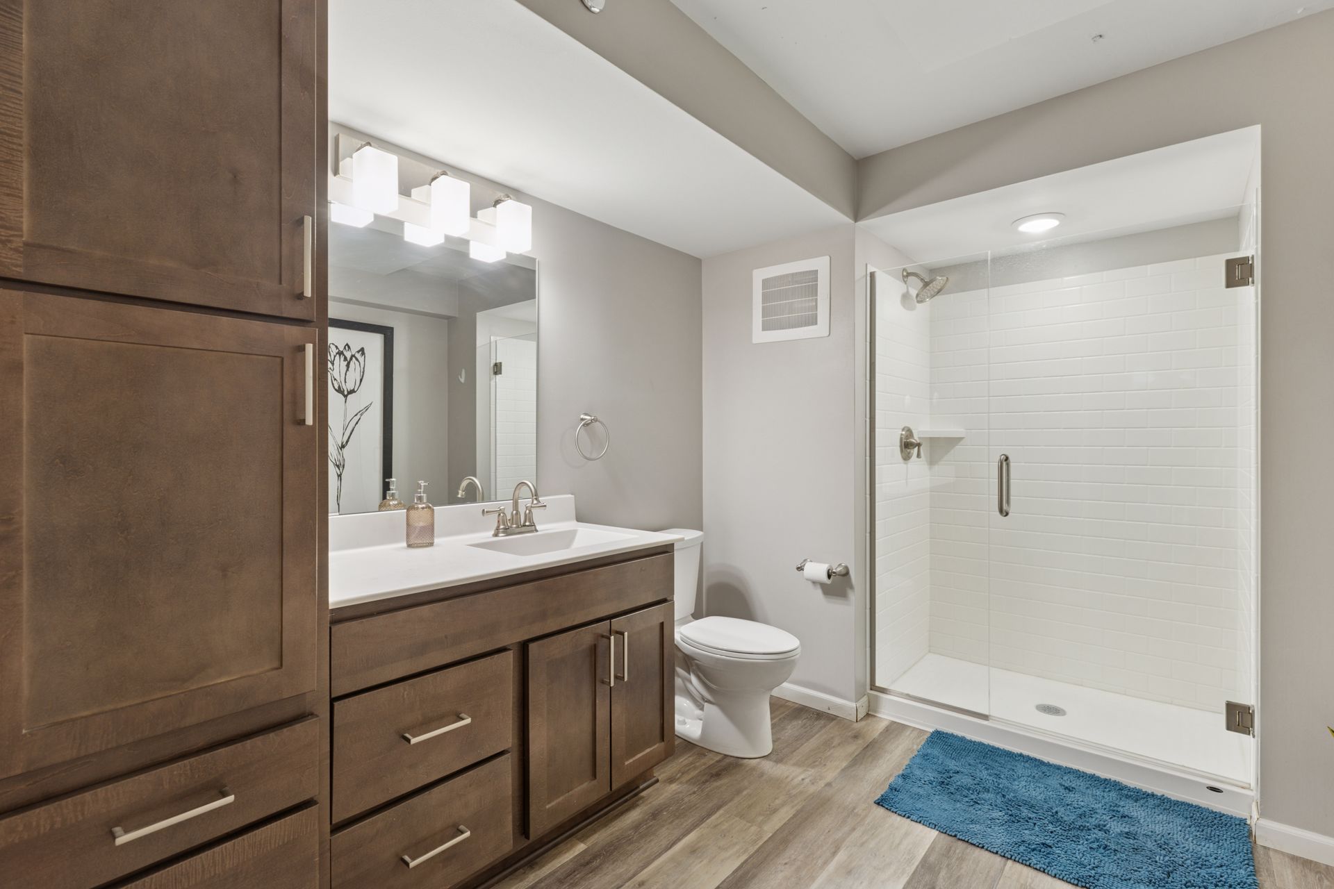 Bathroom with brown cabinets, white countertop, and glass shower door. Blue rug on wood floor at R7 Lofts in St. Paul, MN.