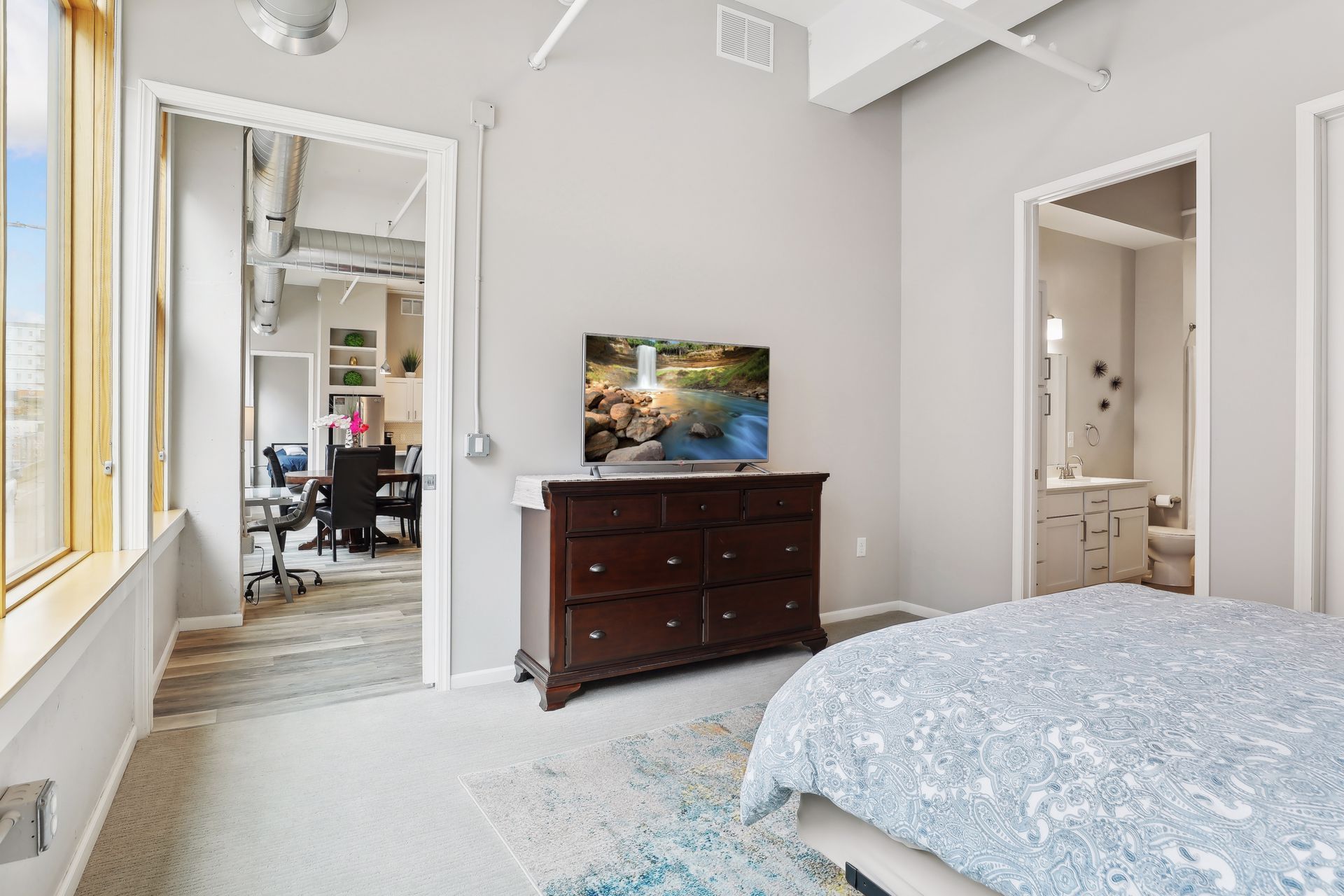 Bedroom with TV on a dark dresser, bed with blue cover, doorway to office, and doorway to bathroom.
