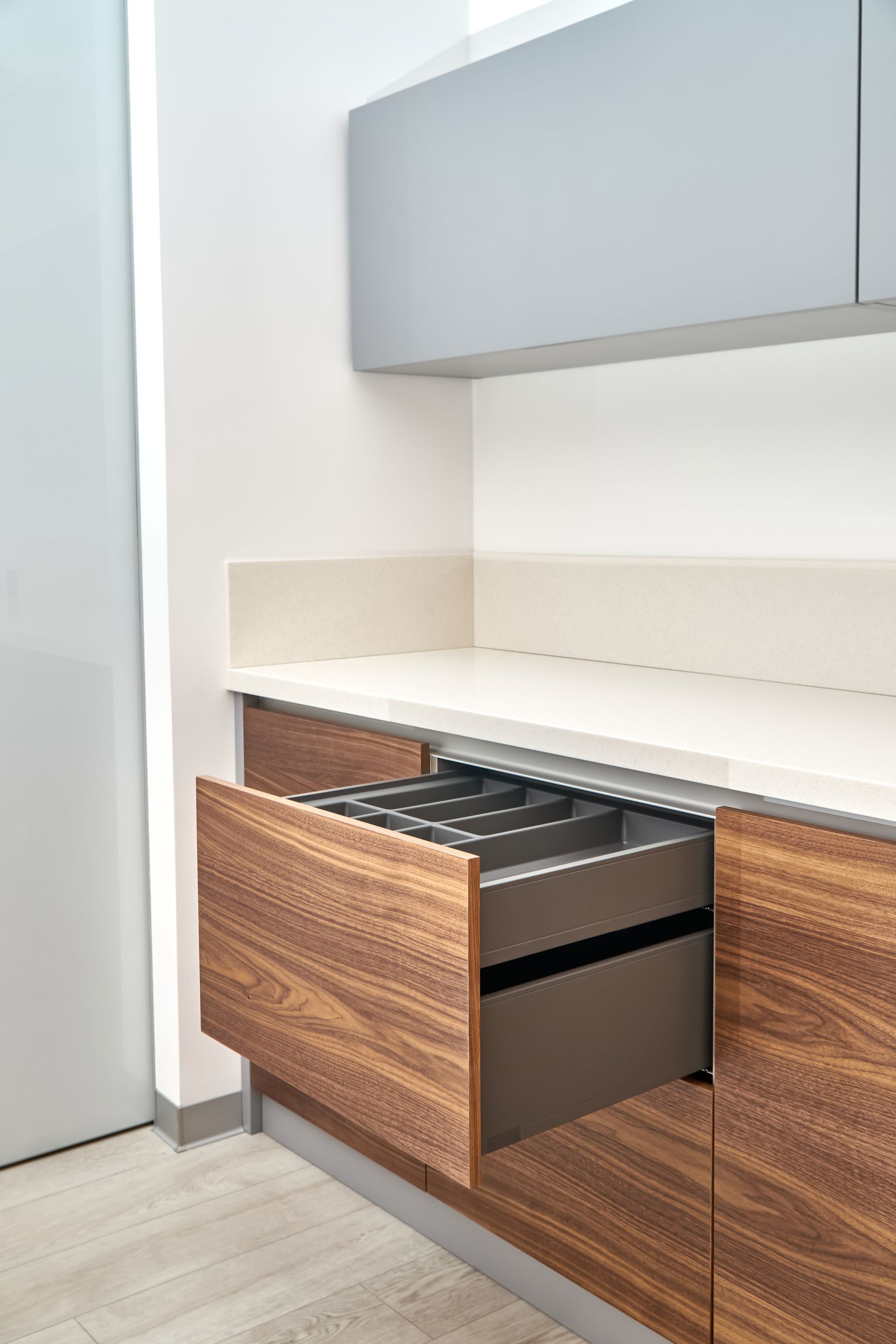 Open kitchen drawer with a silverware organizer, wood grain finish. Gray countertop, white wall, gray upper cabinet.