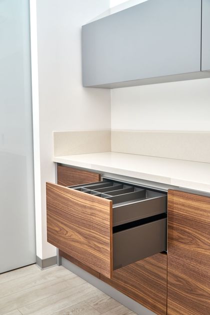 Open kitchen drawer with a silverware organizer, wood grain finish. Gray countertop, white wall, gray upper cabinet. Open kitchen drawer with a silverware organizer, wood grain finish. Gray countertop, white wall, gray upper cabinet.