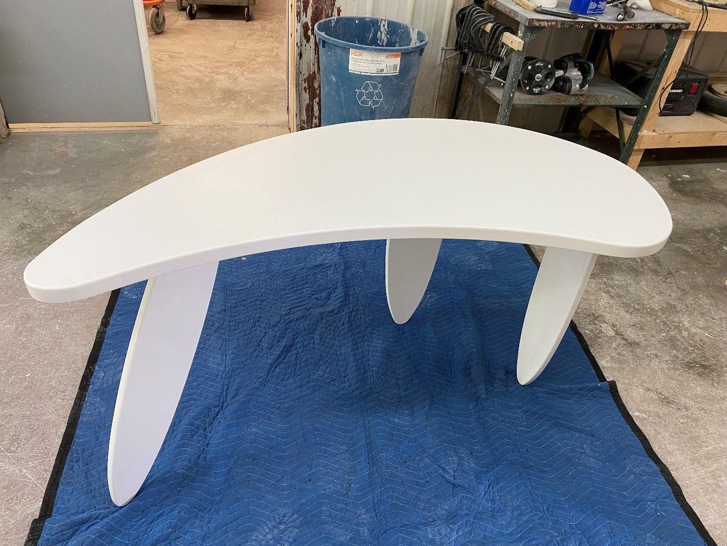 White, curved coffee table with three angled legs, sitting on a blue drop cloth. Workshop in background.