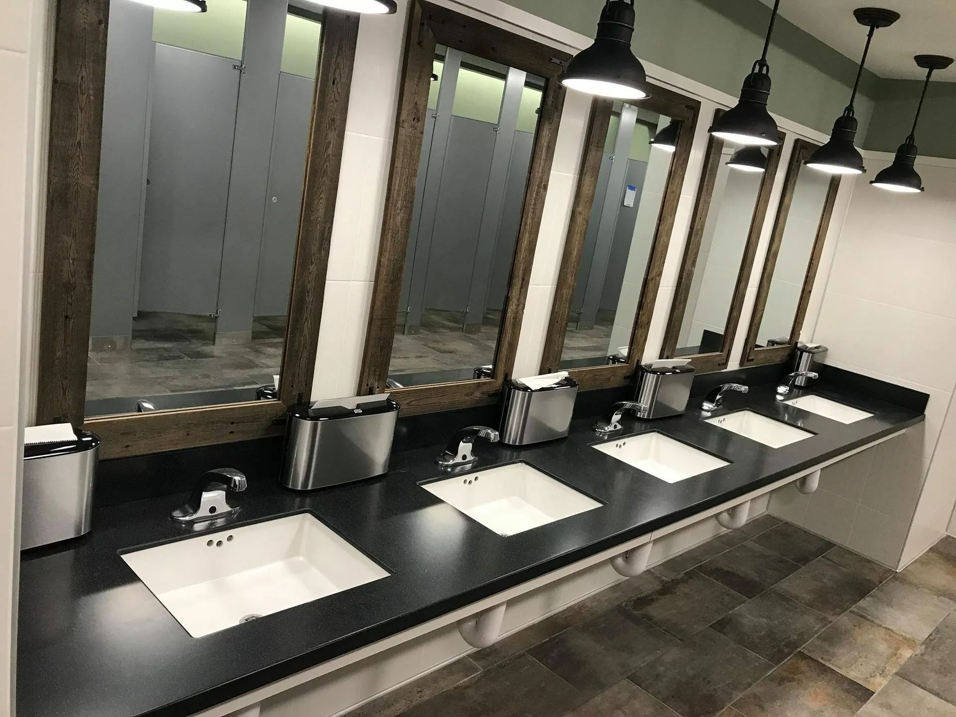Public restroom with five sinks and mirrors, black countertop, gray-tiled floor, and rustic wooden mirror frames.