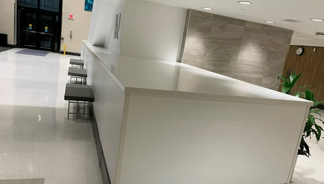 White reception desk with seating, in a modern office lobby. Concrete wall and potted plant visible.