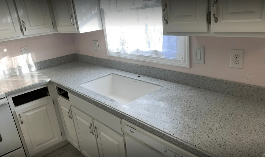 White kitchen with grey speckled countertops, white cabinets, and a sink beneath a window.