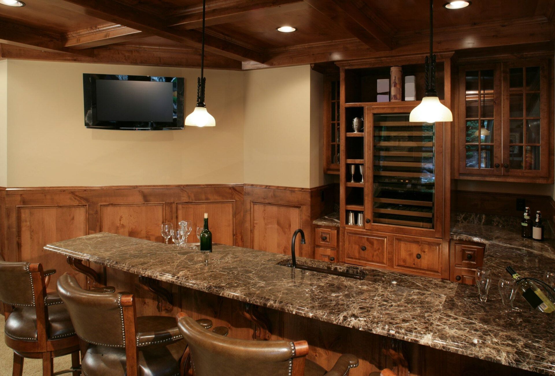 Basement bar with granite countertop, wooden cabinets, and a wine fridge. TV mounted above bar.