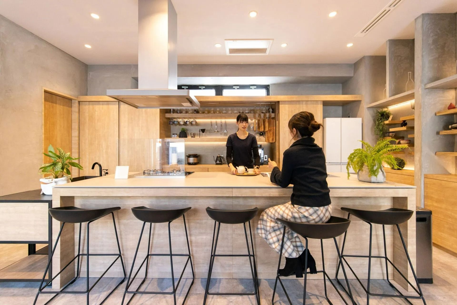 Two women in modern kitchen with island and stools; one seated, other behind the counter.