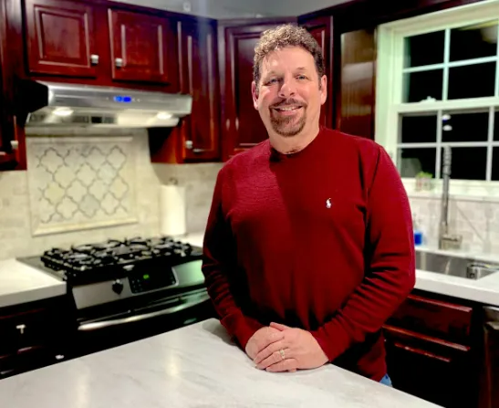 Man in red sweater, smiling, in a kitchen with dark cabinets, white countertops, and stove.