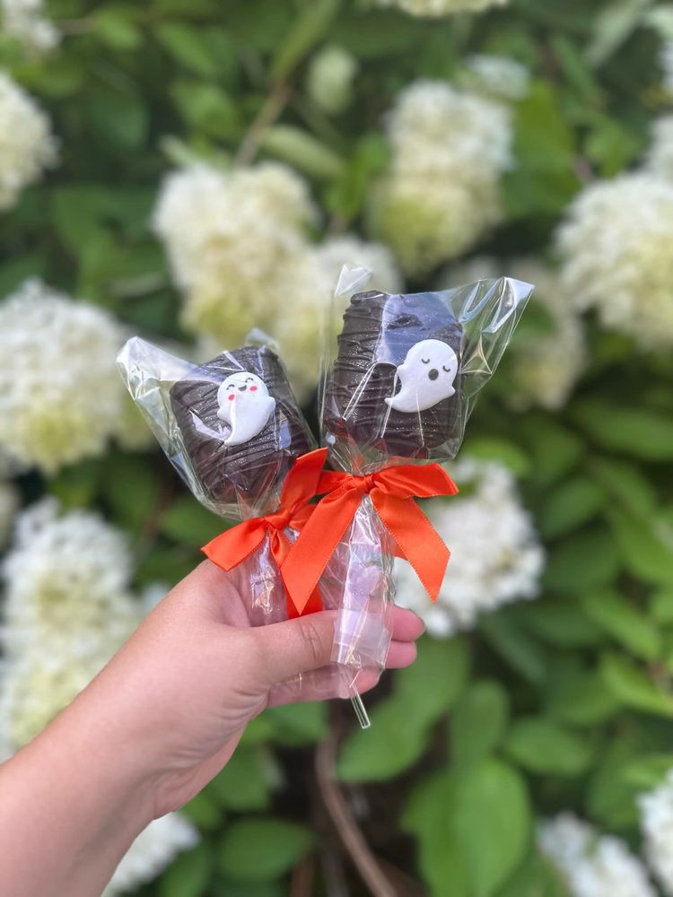 Hand holding two clear ribbon-wrapped treats with orange bows against white flowers and green leaves