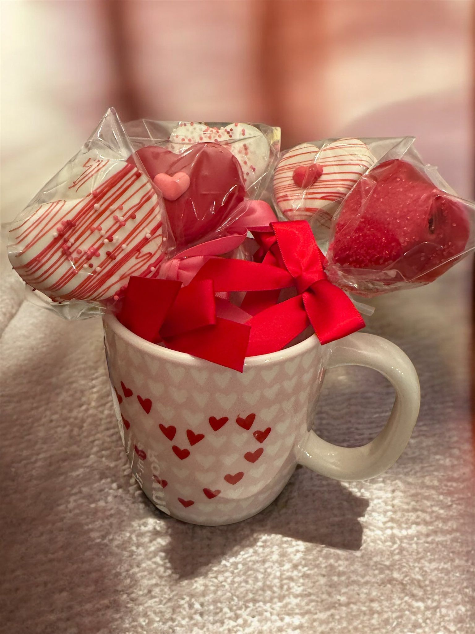 Heart-print mug filled with red ribbon, with wrapped heart-shaped candies behind it on a soft pink surface