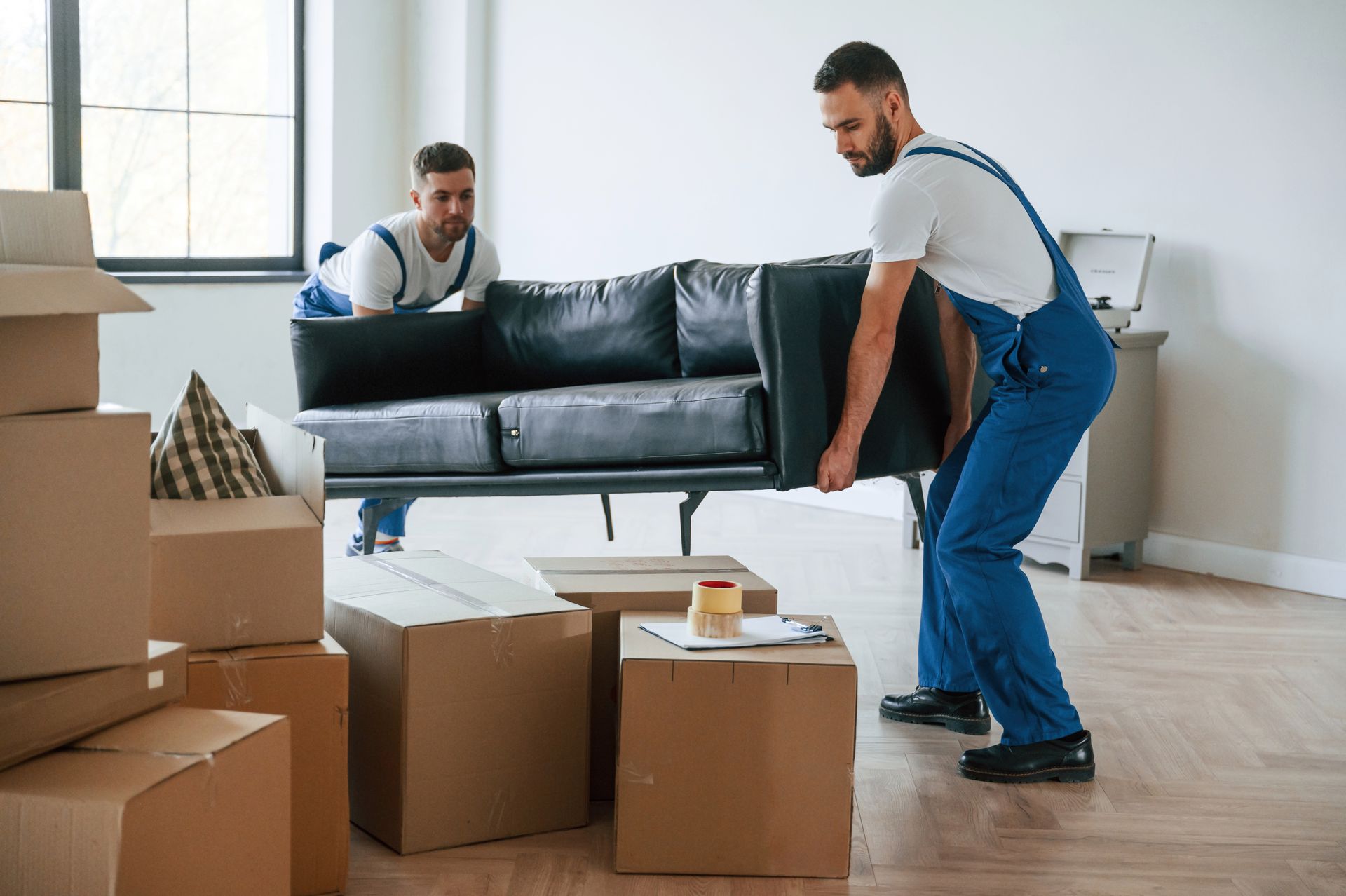 Two movers in blue overalls lift a black sofa amid taped boxes in a bright room.
